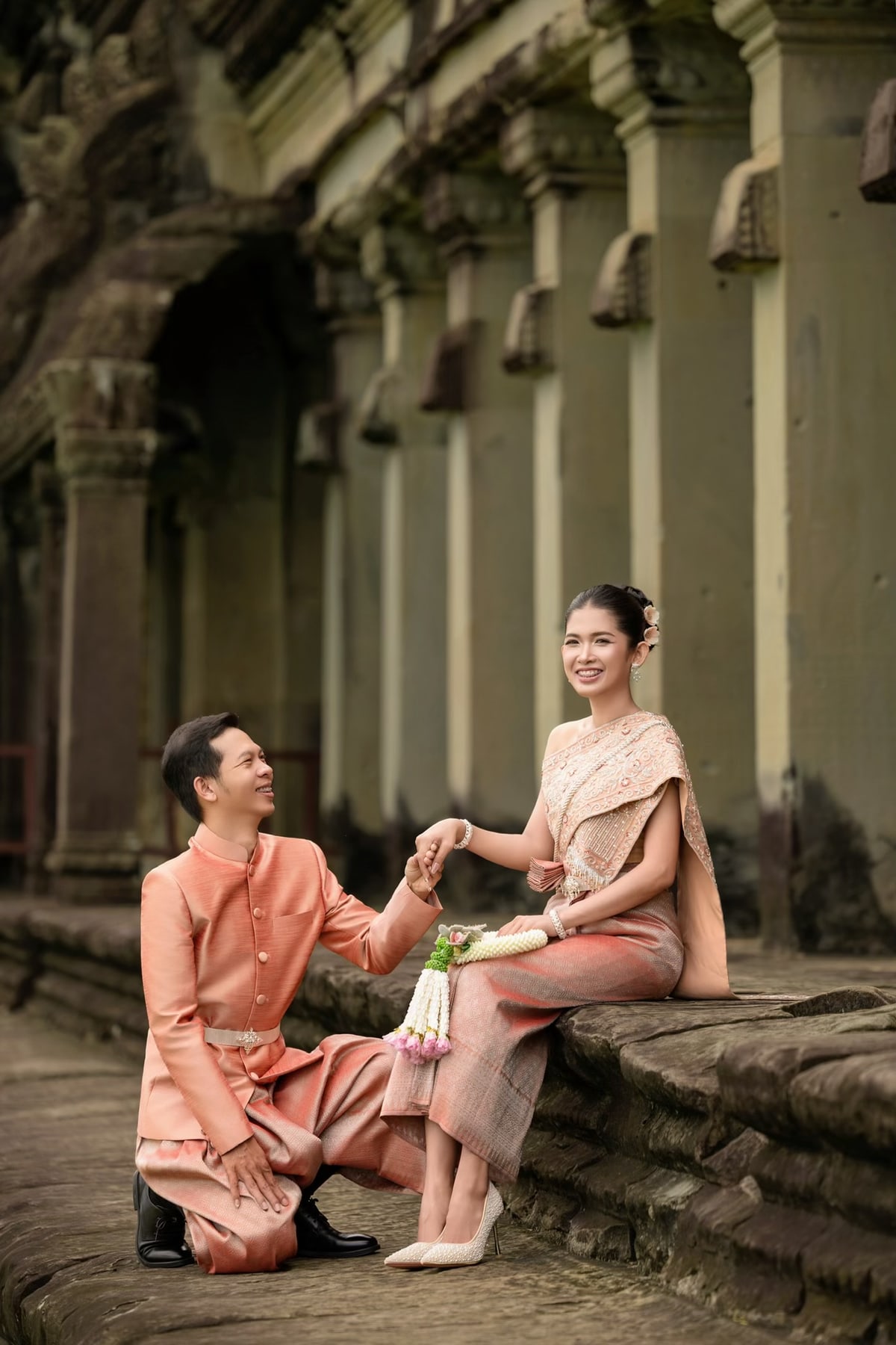 The couple in peach wedding attire beside a stone temple corridor