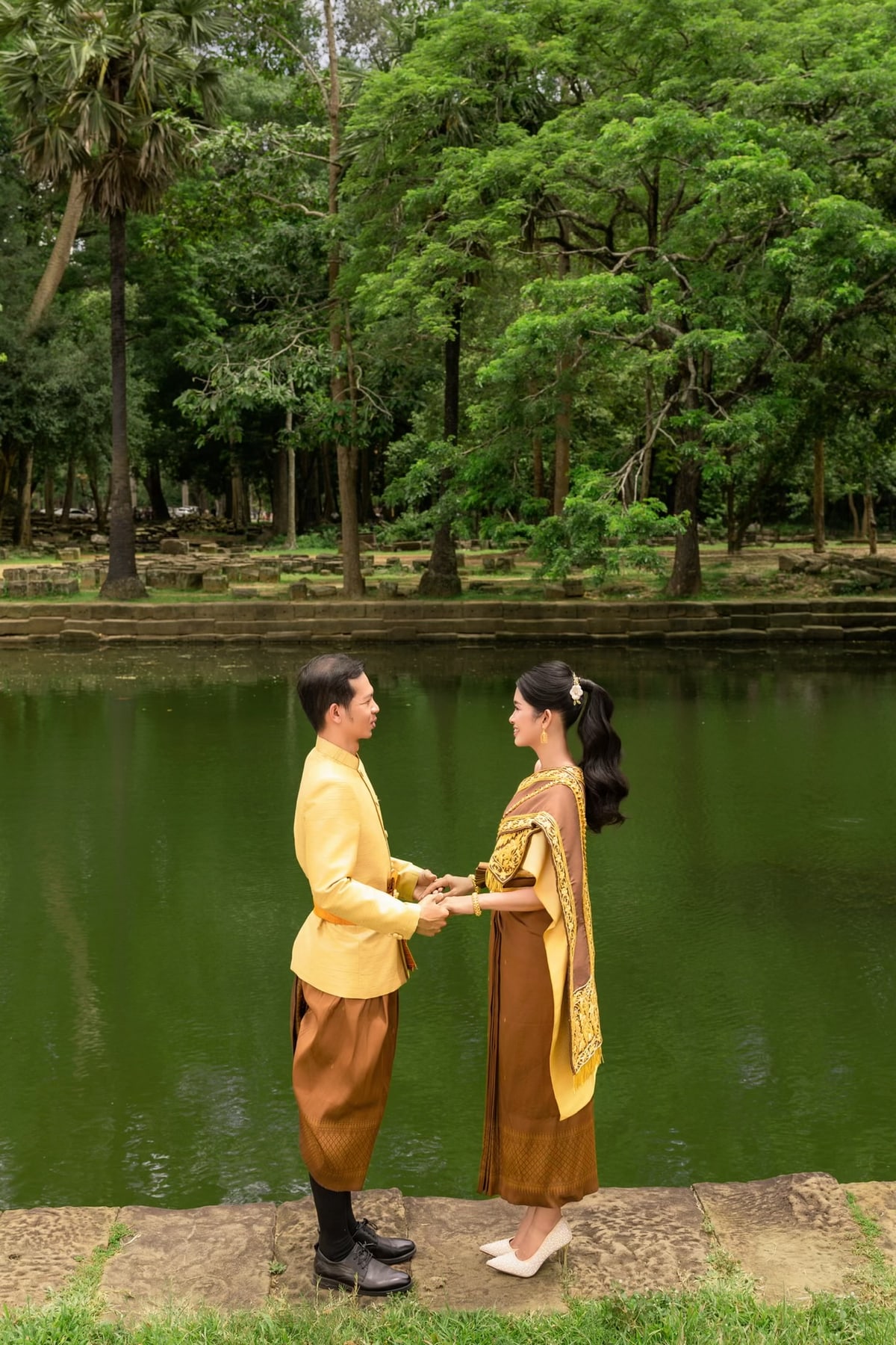 The couple in gold wedding attire holding hands beside a pond